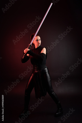 Full length portrait of pretty redhead female model wearing black futuristic scifi leather cloak costume. Standing pose holding lightsaber on dark studio background with shadow moody lighting.