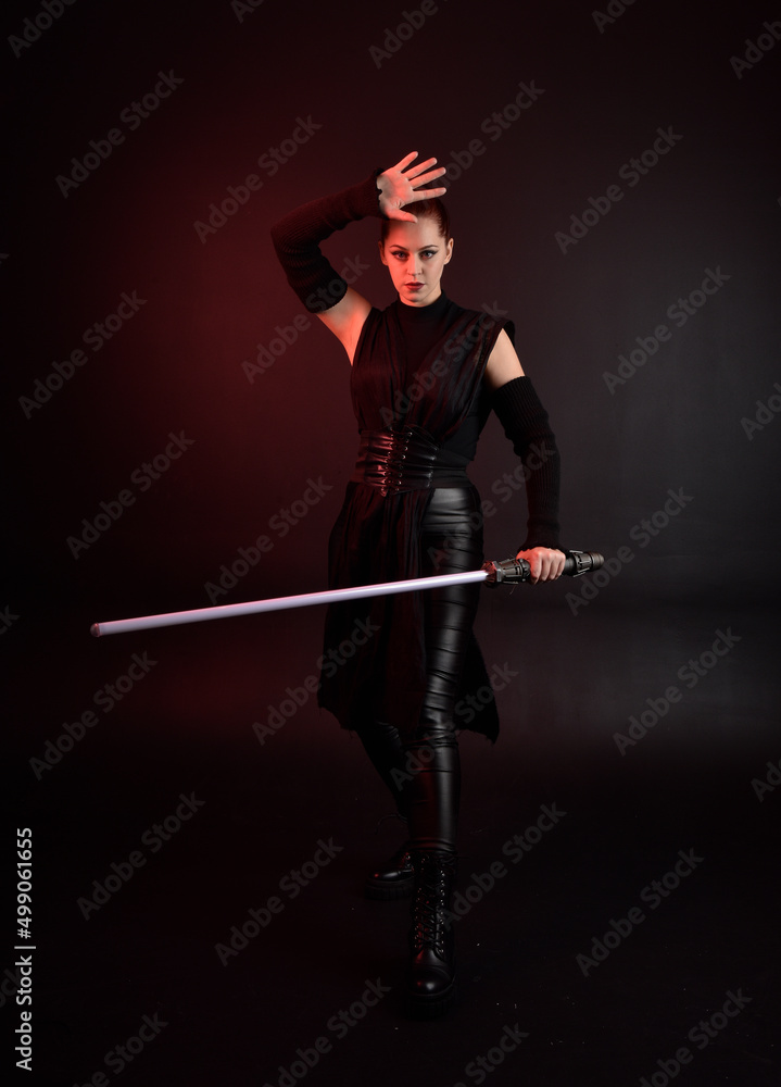 Full length portrait of pretty redhead female model wearing black futuristic scifi leather cloak costume. Standing pose holding lightsaber on dark studio background with shadow moody lighting. Full length portrait of pretty redhead female model wearing black futuristic scifi leather cloak costume. Standing pose holding lightsaber on dark studio background with shadow moody lighting.