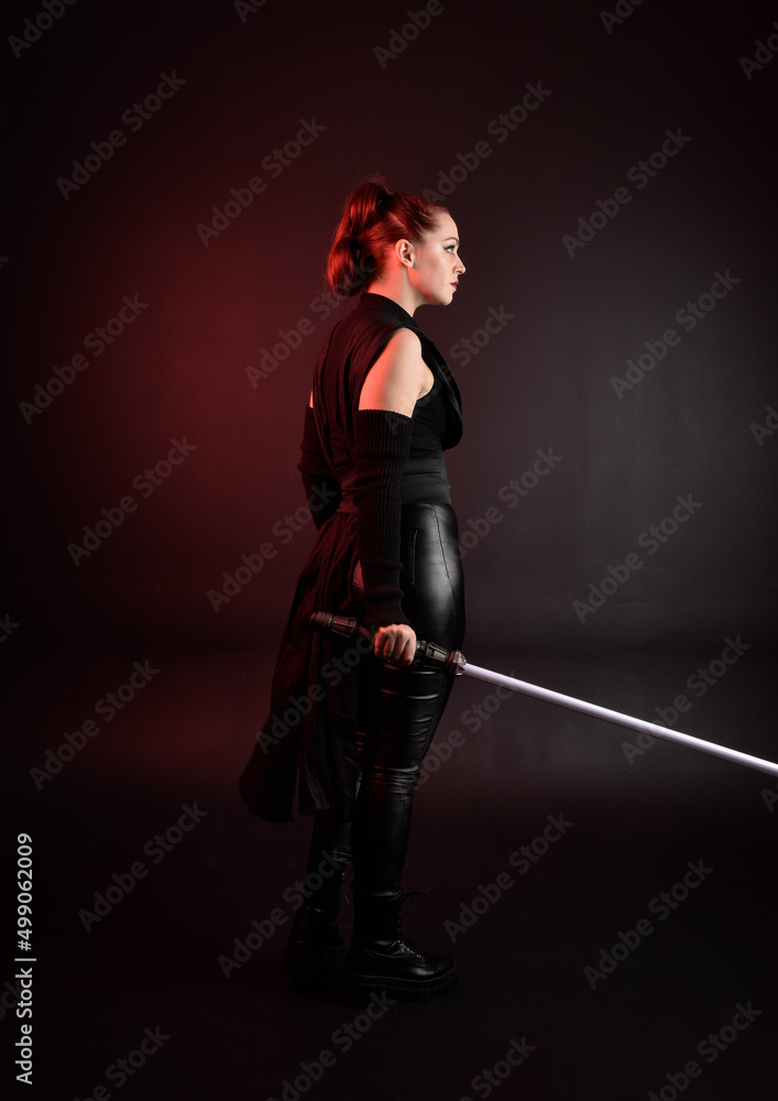 Full length portrait of pretty redhead female model wearing black futuristic scifi leather cloak costume. Standing pose holding lightsaber on dark studio background with shadow moody lighting. Full length portrait of pretty redhead female model wearing black futuristic scifi leather cloak costume. Standing pose holding lightsaber on dark studio background with shadow moody lighting.