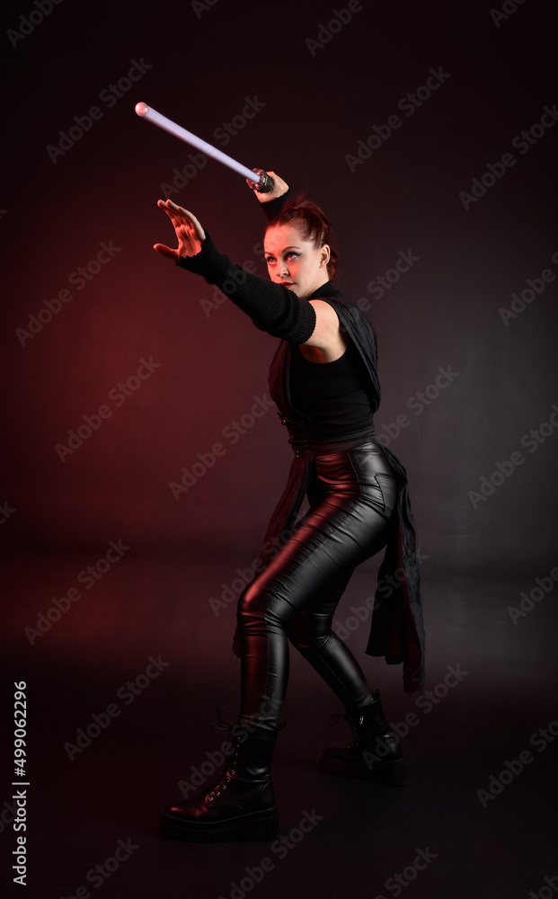 Full length portrait of pretty redhead female model wearing black futuristic scifi leather cloak costume. Standing pose holding lightsaber on dark studio background with shadow moody lighting. Full length portrait of pretty redhead female model wearing black futuristic scifi leather cloak costume. Standing pose holding lightsaber on dark studio background with shadow moody lighting.