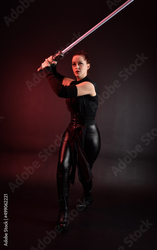 Full length portrait of pretty redhead female model wearing black futuristic scifi leather cloak costume. Standing pose  holding lightsaber on dark studio background with shadow moody lighting.