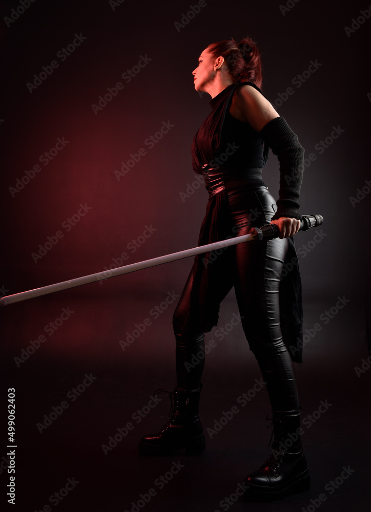 Full length portrait of pretty redhead female model wearing black futuristic scifi leather cloak costume. Standing pose holding lightsaber on dark studio background with shadow moody lighting. Full length portrait of pretty redhead female model wearing black futuristic scifi leather cloak costume. Standing pose holding lightsaber on dark studio background with shadow moody lighting.