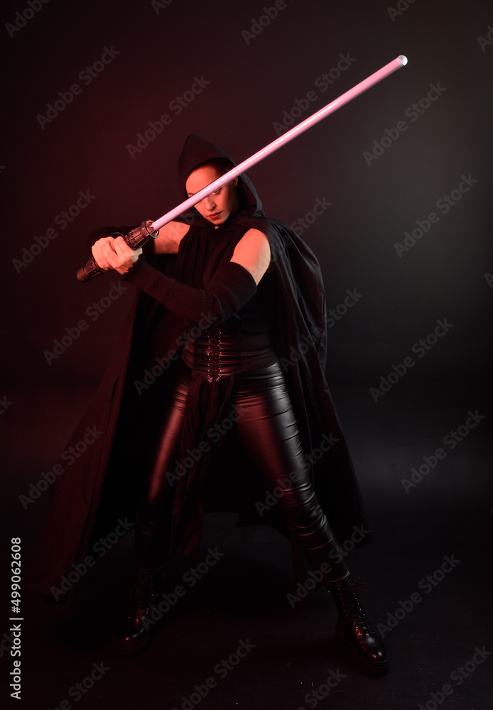 Full length portrait of pretty redhead female model wearing black futuristic scifi leather cloak costume. Standing pose holding lightsaber on dark studio background with shadow moody lighting. Full length portrait of pretty redhead female model wearing black futuristic scifi leather cloak costume. Standing pose holding lightsaber on dark studio background with shadow moody lighting.