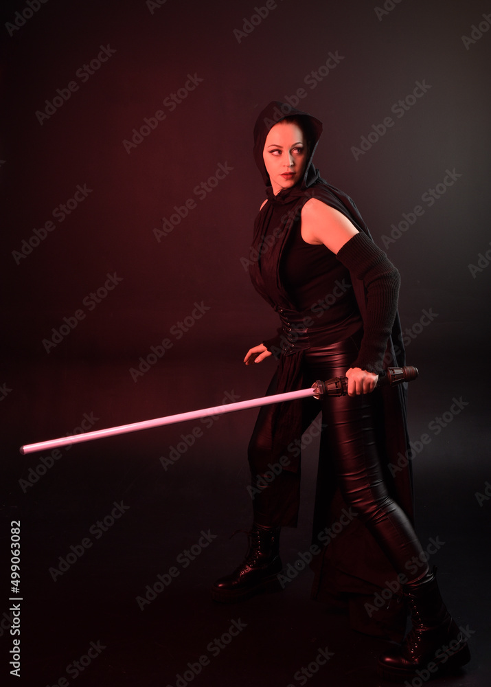 Full length portrait of pretty redhead female model wearing black futuristic scifi leather cloak costume. Standing pose holding lightsaber on dark studio background with shadow moody lighting. Full length portrait of pretty redhead female model wearing black futuristic scifi leather cloak costume. Standing pose holding lightsaber on dark studio background with shadow moody lighting.