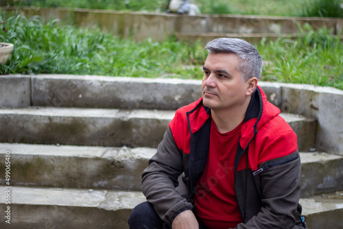 A man in red jacket sits on concrete steps in a garden. 