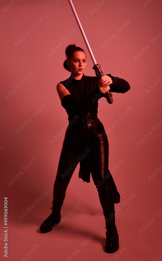 Full length portrait of pretty redhead female model wearing black futuristic scifi leather cloak costume. Standing pose holding lightsaber on red studio background with coloured lighting Full length portrait of pretty redhead female model wearing black futuristic scifi leather cloak costume. Standing pose holding lightsaber on red studio background with coloured lighting