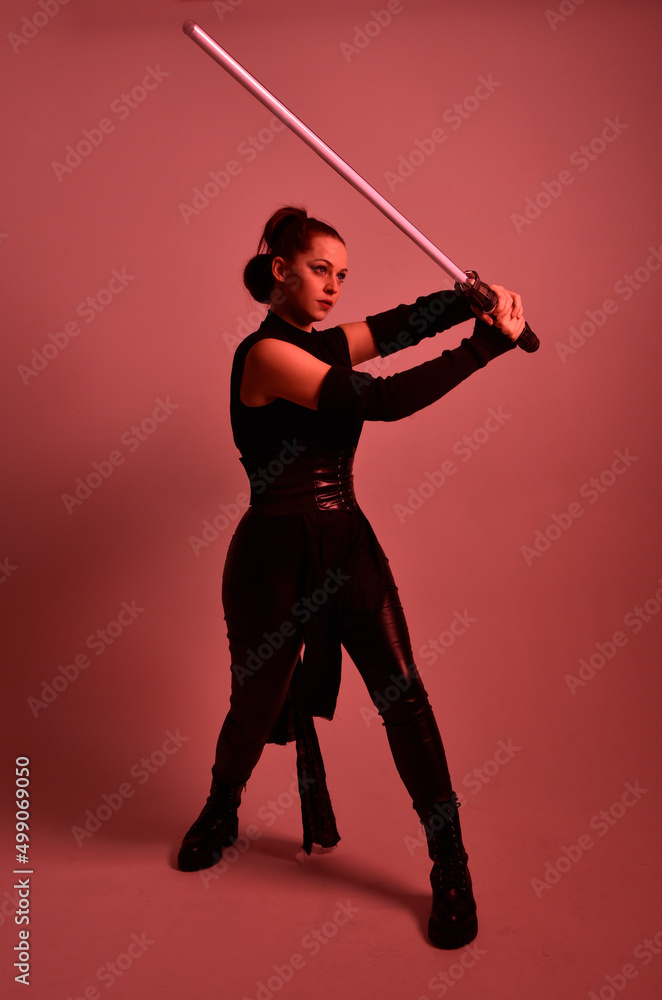 Full length portrait of pretty redhead female model wearing black futuristic scifi leather cloak costume. Standing pose holding lightsaber on red studio background with coloured lighting Full length portrait of pretty redhead female model wearing black futuristic scifi leather cloak costume. Standing pose holding lightsaber on red studio background with coloured lighting