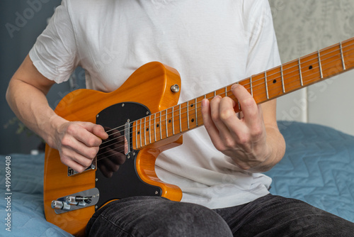 A man in a white T-shirt plays a natural-colored electric guitar sitting on a bed in close-up, selective focus.A male musician plays an electric guitar.Telecaster of natural color.Man playing guitar.  photo