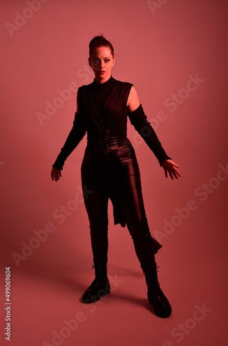 Full length portrait of pretty redhead female model wearing black futuristic scifi leather cloak costume. Standing pose  holding lightsaber on red studio background with coloured lighting © faestock