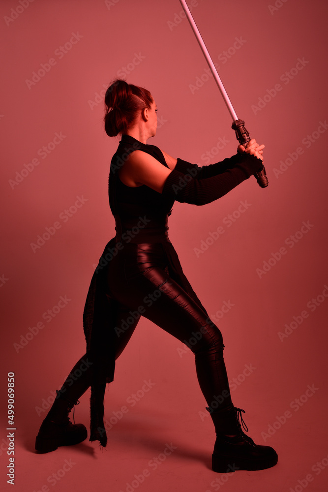 Full length portrait of pretty redhead female model wearing black futuristic scifi leather cloak costume. Standing pose holding lightsaber on red studio background with coloured lighting Full length portrait of pretty redhead female model wearing black futuristic scifi leather cloak costume. Standing pose holding lightsaber on red studio background with coloured lighting
