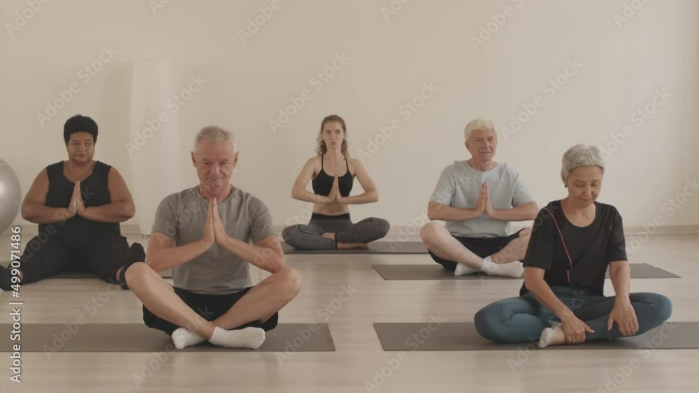 Stab shot of group of four multiethnic active seniors and young female yoga coach sitting on yoga mats in lotus pose meditating with praying hands Stab shot of group of four multiethnic active seniors and young female yoga coach sitting on yoga mats in lotus pose meditating with praying hands