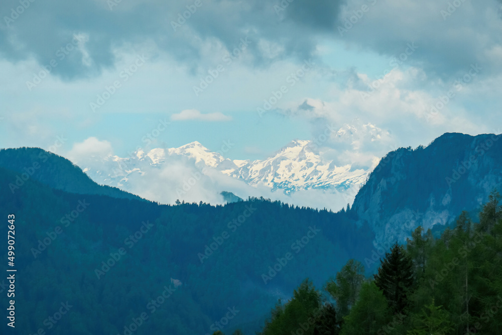 Scenic view on the snow capped and cloud covered mountains of Julian Alps and Kamnik Savinja Alps in Carinthia, border Austria and Slovenia. Dense green forest in spring in the valley. Mountaineering