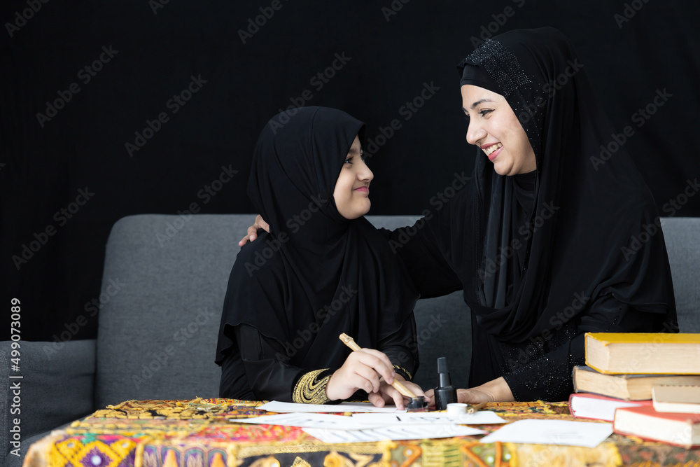 muslim mother with her daughter writing Arabic text with bamboo pens and black ink on paper, Arabic letters mean the name of Muslim god muslim mother with her daughter writing Arabic text with bamboo pens and black ink on paper, Arabic letters mean the name of Muslim god
