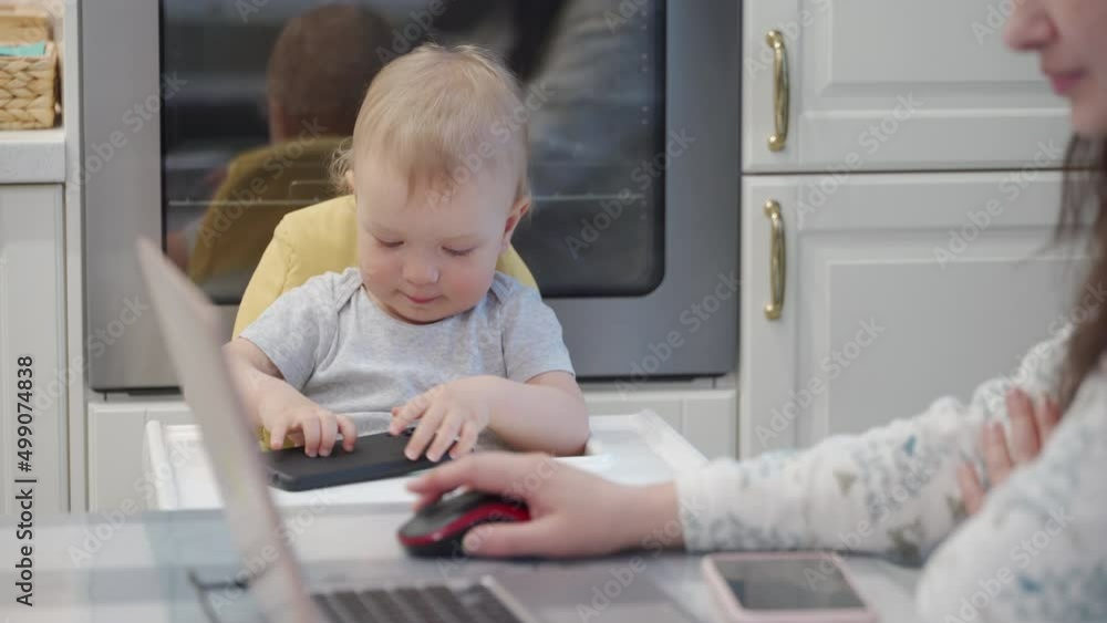 Multitasking mom with small child working remote, woman using laptop, baby boy playing with mobile phone. Mother working and parenting. High quality 4k footage Multitasking mom with small child working remote, woman using laptop, baby boy playing with mobile phone. Mother working and parenting. High quality 4k footage
