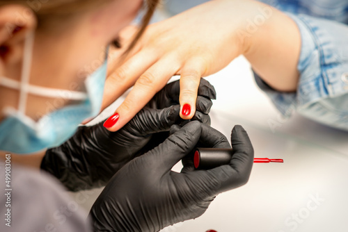 Manicure varnish painting. Close-up of a manicure master wearing rubber black gloves and a protective mask applying red varnish on a female fingernail in the beauty salon