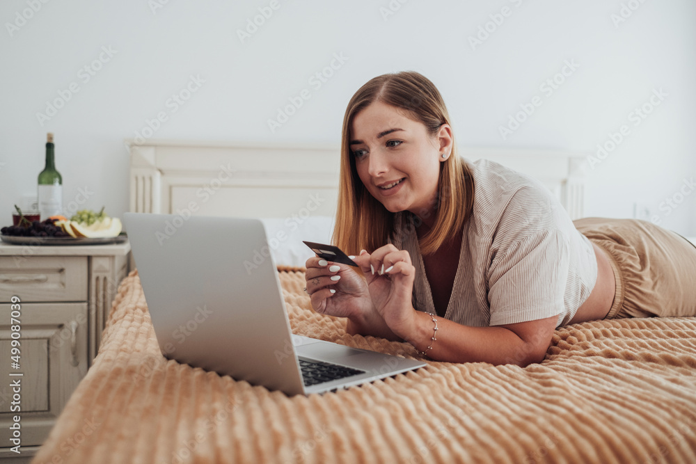 Caucasian Young Woman Making Online Shopping Through Laptop While Laying on the Bed of Hotel Room Caucasian Young Woman Making Online Shopping Through Laptop While Laying on the Bed of Hotel Room