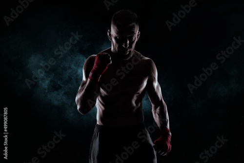 Kickboxer in red bandages poses against a background of smoke. The concept of mixed martial arts. © andy_gin