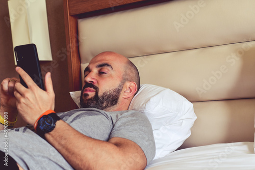 Handsome bearded man in pajamas using a cellphone while lying in bed after waking up in the morning