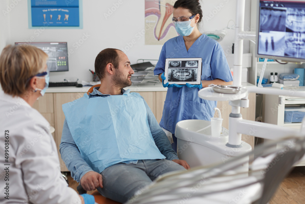 Dentist assistant with face mask holding tabet computer explaining teeth radiography to sick patient during dentistry consultation in stomatological office room. Team working at caries treatment Dentist assistant with face mask holding tabet computer explaining teeth radiography to sick patient during dentistry consultation in stomatological office room. Team working at caries treatment