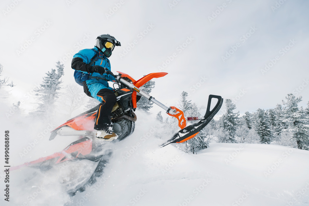 Jumping Snowbike rider in mountain valley in beautiful snow powder. Modify motorcycle with single ski in the front and special snowmobile-style track in the back instead of wheels Jumping Snowbike rider in mountain valley in beautiful snow powder. Modify motorcycle with single ski in the front and special snowmobile-style track in the back instead of wheels