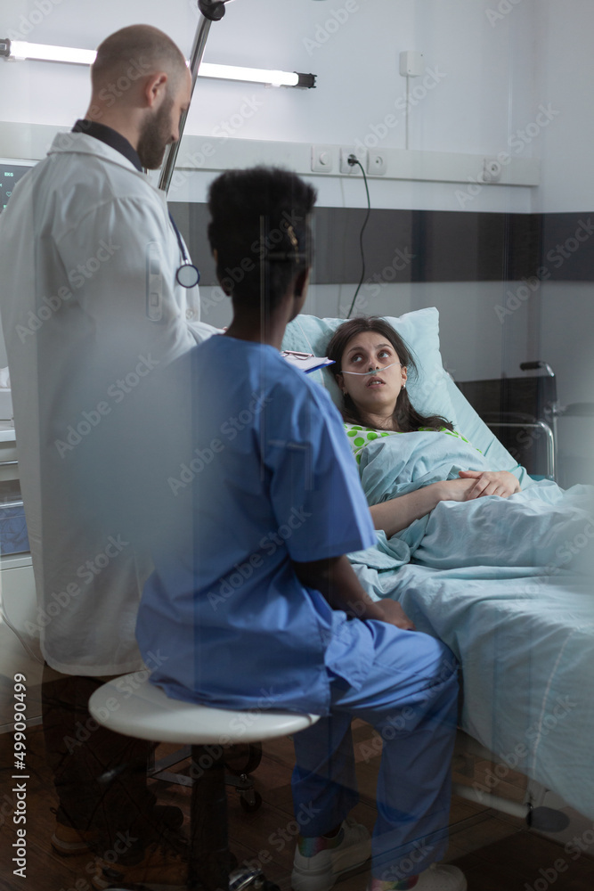 Through the window view of doctor with clipoboard and nurse in private ward consulting recovering patient after medical procedure. Patient with nasal cannula looking at health care professionals. Through the window view of doctor with clipoboard and nurse in private ward consulting recovering patient after medical procedure. Patient with nasal cannula looking at health care professionals.