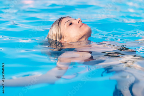 A young beautiful blonde woman in a black swimsuit swims on her back with her eyes closed in the turquoise water of the pool.Summer concept.