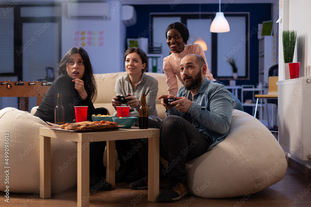 Smiling people playing video games online on tv console, using controller to win at office part after work. Colleagues having fun with game, eating pizza and drinking alcoholic beer.