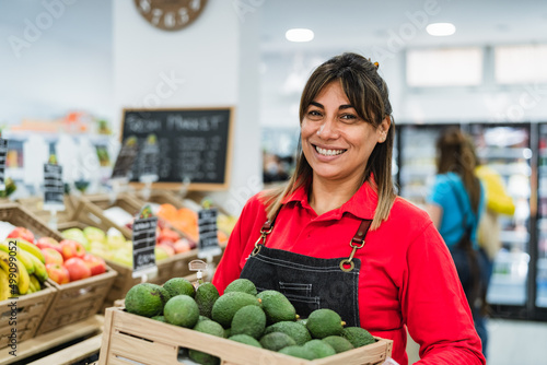 Latin woman working in supermarket holding a box containing fresh avocados