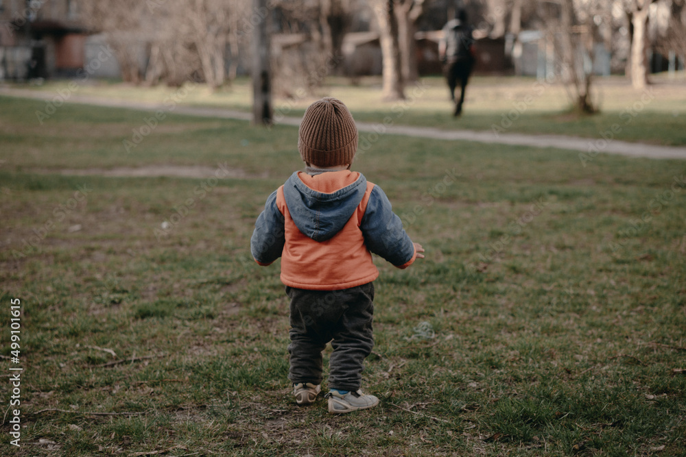 A little boy stands with his back in an orange jacket on green grass A little boy stands with his back in an orange jacket on green grass