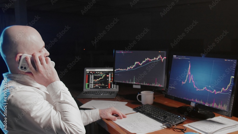Side view of a man sitting at a computer and looking at an online stock market chart showing bitcoin currencies. A man is talking on the phone. In real time. Cryptocurrency. Investors