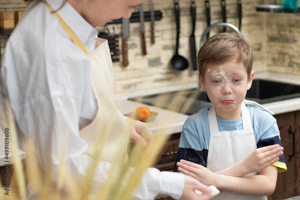 Mom and son in aprons play with flour while cooking at home in the kitchen against the background of kitchen utensils. Selective focus. Portrait. Close-up Mom and son in aprons play with flour while cooking at home in the kitchen against the background of kitchen utensils. Selective focus. Portrait. Close-up