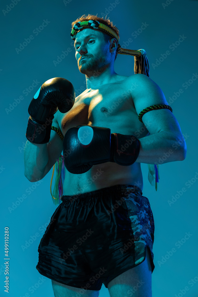 Portrait of muscled man, thai boxer in sports uniform posing isolated on blue studio background in neon. Sport, muay thai, competition, fight club concept. Bottom view Portrait of muscled man, thai boxer in sports uniform posing isolated on blue studio background in neon. Sport, muay thai, competition, fight club concept. Bottom view
