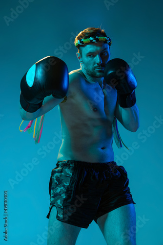 Half-length portrait of muscled man, thai boxer in sports uniform posing isolated on blue studio background in neon. Sport, muay thai, competition, fight club concept