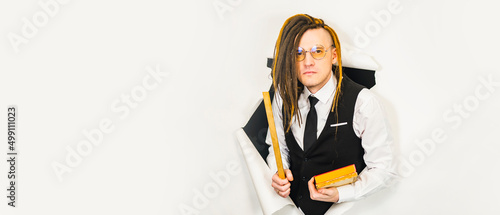 Young teacher in glasses with ruler and books in hole of white background. Male pedagogue with modern hairstyle, dreadlocks looking at camera in studio.