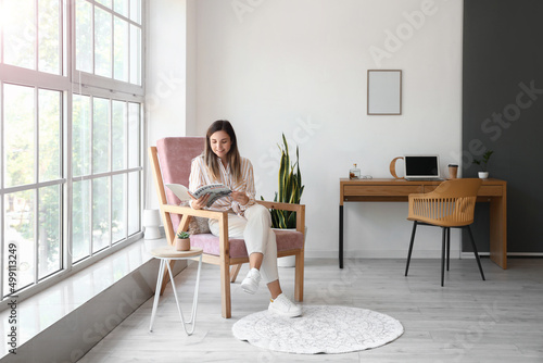 Young woman with magazine sitting in comfortable armchair at home