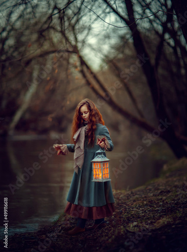 Magical girl with lantern in her hand standing near the river in the warm colored park