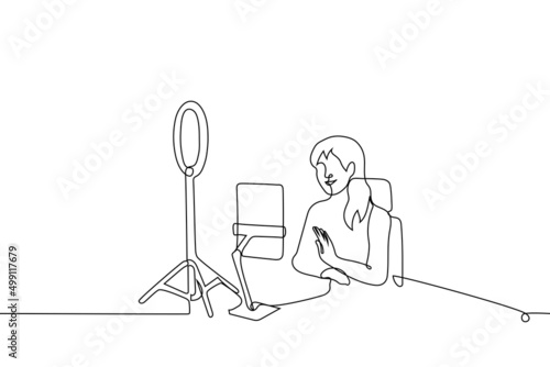 female blogger smiles and waves at the camera of tablet on tripod, it is illuminated by round lamp for video light - one line drawing vector. concept of online streaming, recording video lesson, q&a