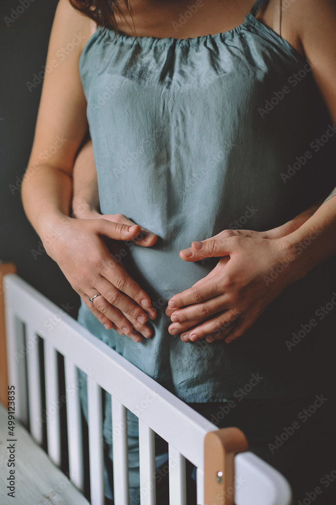 Pregnant woman in blue pajamas and her husband standing near crib and holding their hands on  pregnant belly. The concept of happy motherhood and parenthood