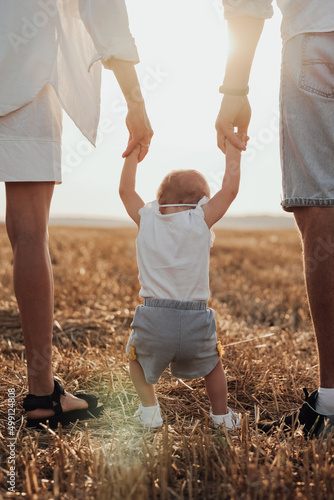 Close Up Back View of Mom and Dad Helping Their Child Making First Steps, Young Family Walking on the Field at Sunset