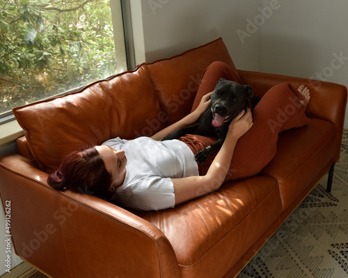 full length portrait o red haired girl wearing casual clothes, relaxing on comfortable leather couch in living room home setting.