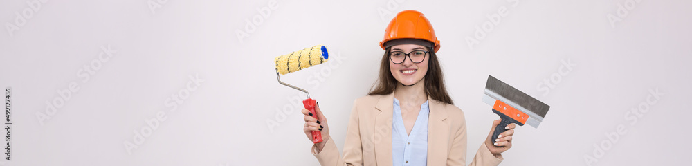 Girl engineer in an orange construction helmet with plastering painting tools in her hands on a white background. Girl engineer in an orange construction helmet with plastering painting tools in her hands on a white background.