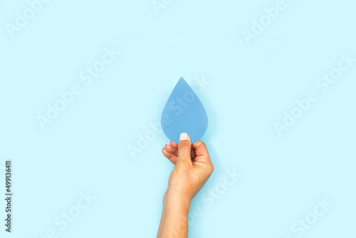 Mano de mujer sosteniendo una gota de agua de papel sobre un fodo celeste liso y aislado. Visa de frente y de cerca. Copy space photo