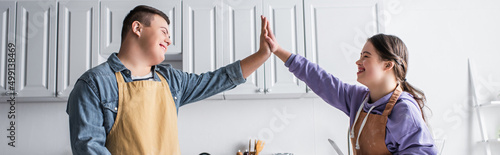 Cheerful friends with down syndrome in aprons giving high five in kitchen, banner. photo