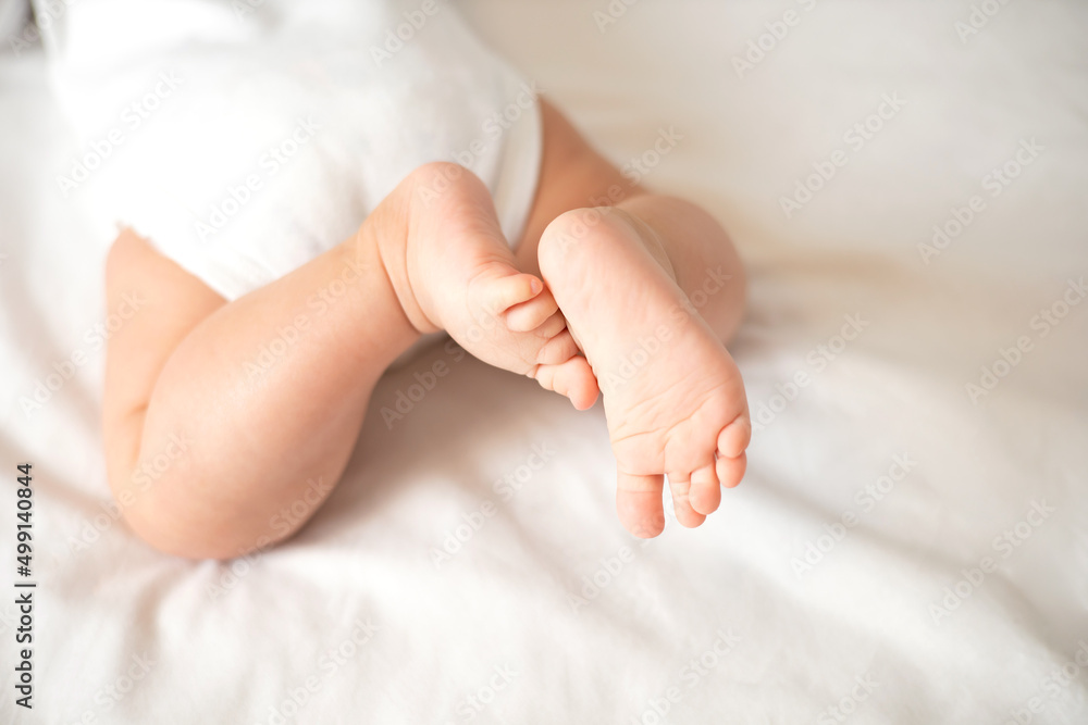The legs of the baby in a white bodysuit on the white background of the bed. Light colors. Lifestyle.