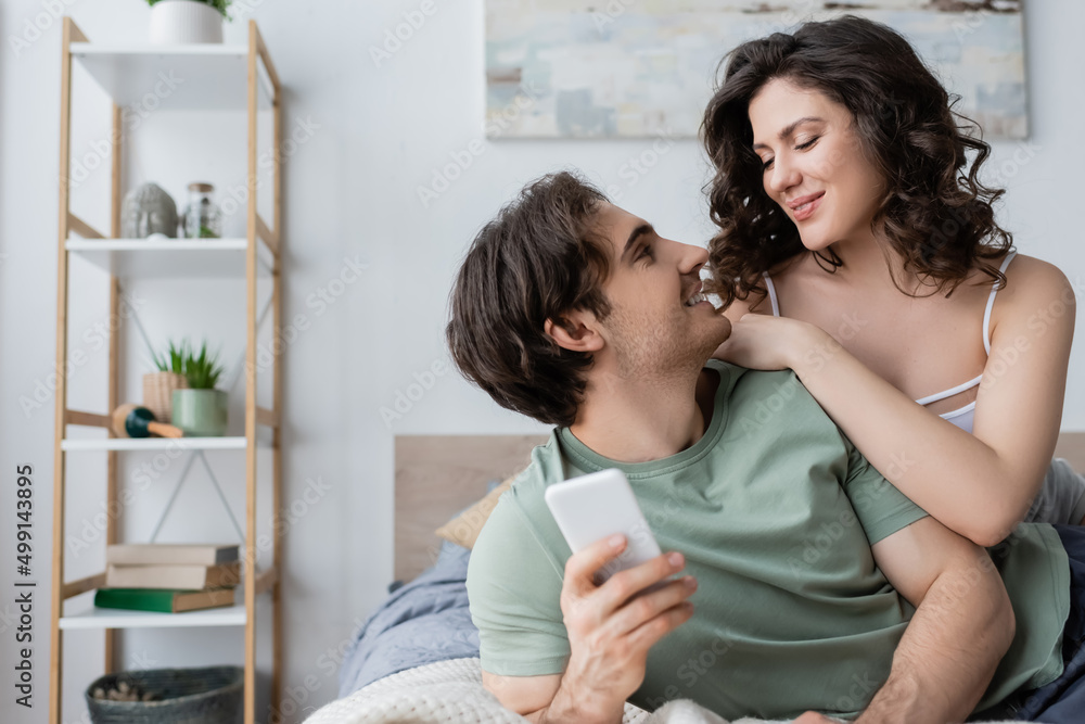 young man holding smartphone and looking at girlfriend in bedroom.