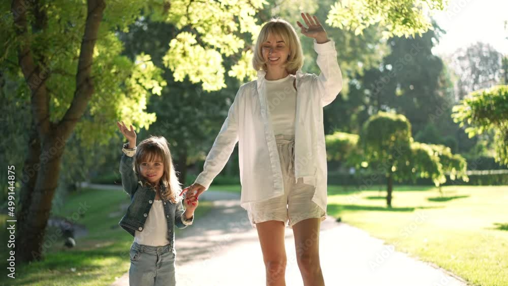 Relaxed excited mother and daughter standing in sunrays smiling waving looking at camera. Caucasian beautiful woman and cute girl posing in sunbeam in slow motion outdoors Relaxed excited mother and daughter standing in sunrays smiling waving looking at camera. Caucasian beautiful woman and cute girl posing in sunbeam in slow motion outdoors