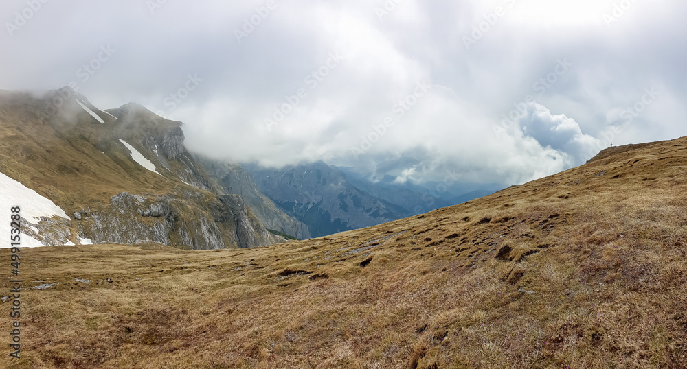 Scenic hiking trail over an alpine meadow in the cloud covered mountain peaks of the Hochschwab Region, Upper Styria, Austria. Rainy spring day in the Alps, Europe. Weather change high altitude. Wind