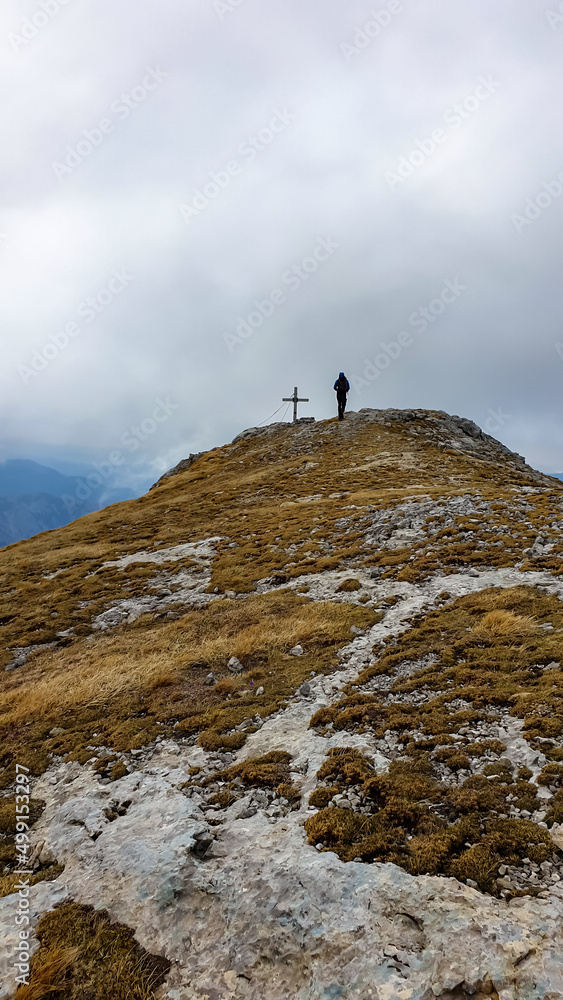 Male person reaching the cloud covered summit cross of mount Zinken in the Hochschwab Region in Upper Styria, Austria. Dry alpine meadows in beautiful Alps in Europe. Freedom vibe. Rainy dark clouds