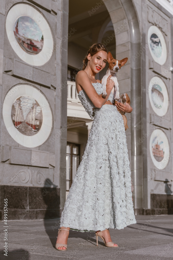 Naklejka premium An incredibly beautiful woman with a dog laughs. She is dressed in a gently turquoise taffeta dress. Luxurious hairstyle and red lipstick make woman bright and memorable 
