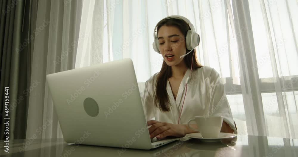 Pretty asian girl in white robe and headphones using laptop computer, having virtual online video conference and talking to friends in morning 4k footage lens flare Pretty asian girl in white robe and headphones using laptop computer, having virtual online video conference and talking to friends in morning 4k footage lens flare